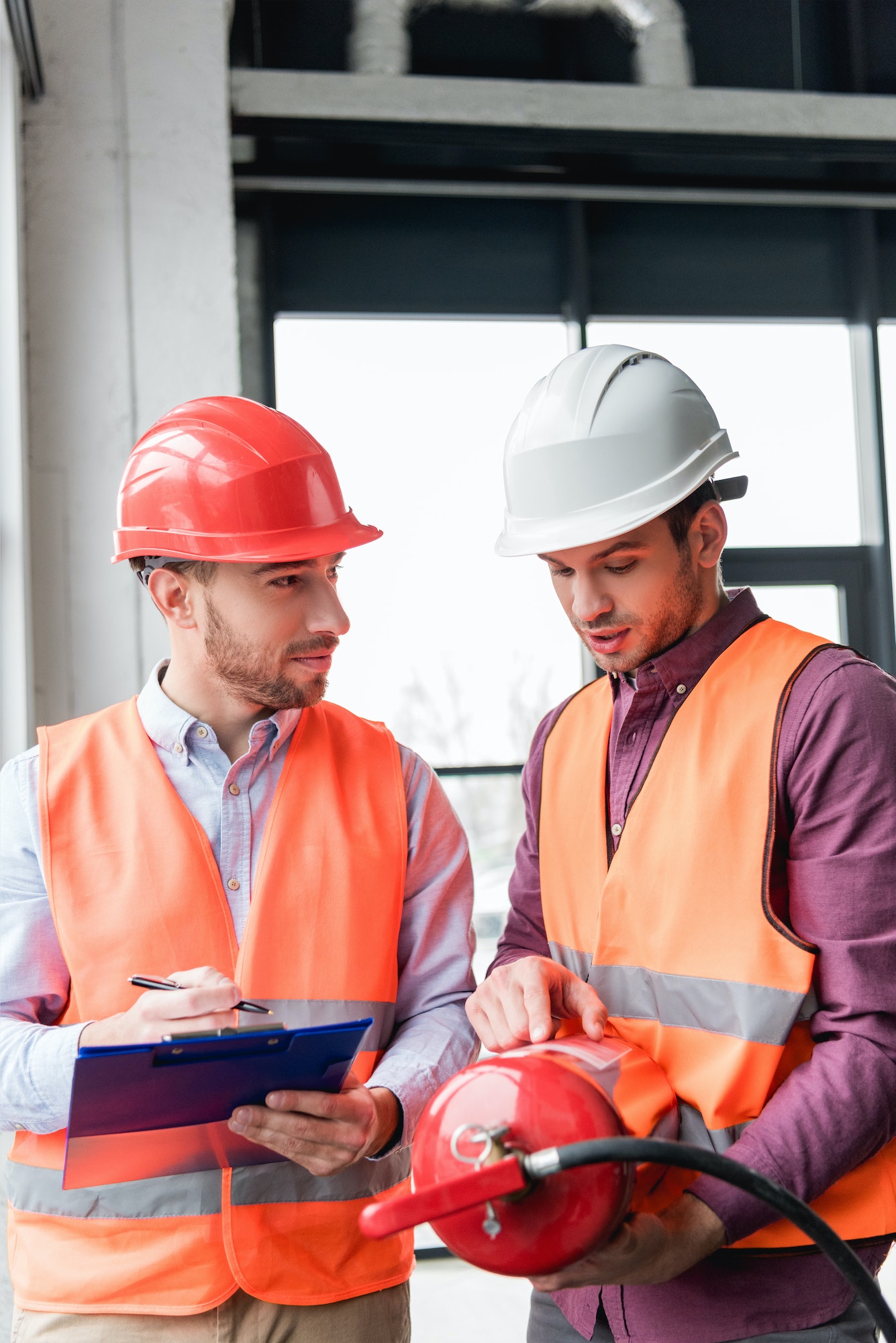 fireman in helmet looking at coworker pointing with finger while holding red extinguisher